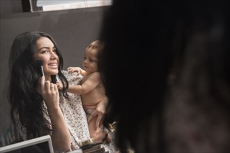 Caucasian mother holding baby son and applying makeup in mirror