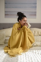 Hispanic woman sitting on bed wrapped in blanket drinking coffee