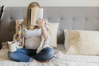 Caucasian expectant mother sitting on bed reading book and drinking coffee
