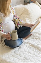 Caucasian expectant mother sitting on bed reading book and drinking coffee