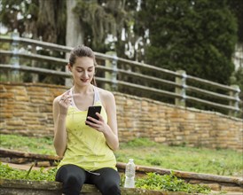 Smiling woman in sport clothing sitting on wall and looking at smart phone