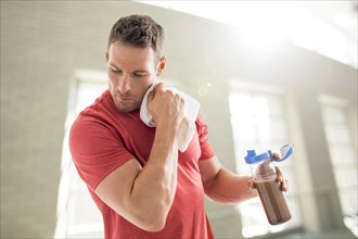 Man holding protein shake and rubbing face with towel in gym