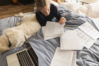 Teenage girl (16-17) lying on bed with dog and doing homework