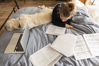 Teenage girl (16-17) lying on bed with dog and doing homework