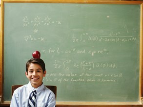 Portrait of boy (12-13) with apple on head sitting in front of chalkboard