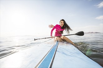 Smiling woman in pink swimsuit sitting on paddleboard and splashing water