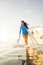 Woman in blue swimsuit carrying paddleboard in lake at sunset