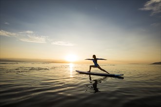 Woman performing Virabhadrasana II (Warrior II Pose) on paddleboard at sunset