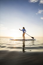 Woman in blue swimsuit paddleboarding on lake at sunset