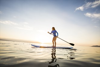 Woman in blue swimsuit paddleboarding on lake at sunset