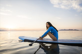 Portrait of woman in blue swimsuit sitting on paddleboard on lake at sunset