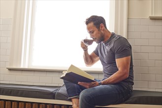 Man reading book and drinking tea by window at home