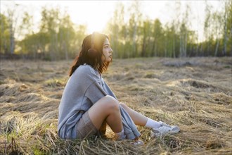Side view of woman listening to music while sitting on grass in meadow at sunset