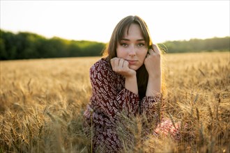 Portrait of young woman looking at camera while crouching in field at sunset