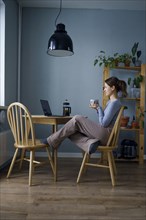Woman holding mug and looking at laptop at home