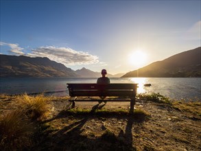 Rear view of woman sitting on bench, facing Lake Wakatipu at sunset