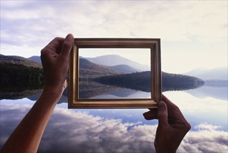 Woman's hands holding picture frame up to calm Lake Placid and hills, Lake Placid, New York, USA