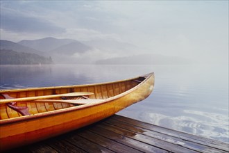 Wooden canoe on dock by calm Lake Placid covered with mist, Lake Placid, New York, USA