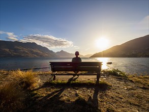 Rear view of woman sitting on bench, facing Lake Wakatipu at sunset, Queenstown, Otago, New Zealand