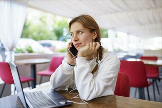 Woman with smart phone, headphones and laptop at cafe table, Omsk, , Russia