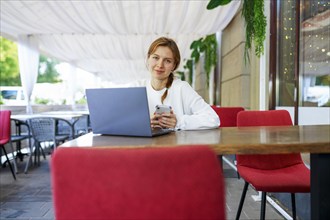 Portrait of smiling woman with smart phone and laptop at cafe table, Omsk, , Russia