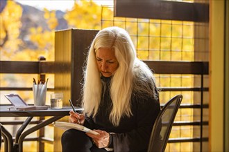 Woman with long white hair painting watercolors on deck in autumn, Springdale, Utah, USA