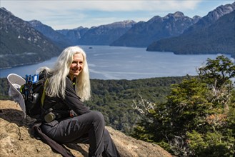 Portrait of smiling female hiker with long white hair sitting on rock above mountain lakes,