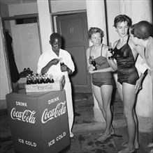 Swim Coca Cola. An African boy opens and distributes bottles of Coca Cola to a group of European
