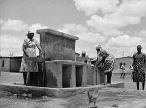 Wash place. Women wearing headscarves and patterned dresses wash bowls and collect water from taps
