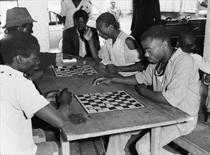Draughts players. A group of men sit on benches around a table to play draughts. An original