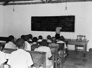 Kenyan school room. A classroom of children sit in rows behind desks listening to a teacher who is