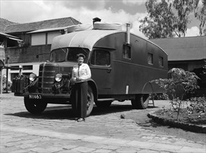 The Price's caravan. A woman stands with her hands clasped beside an old-fashioned caravan in the