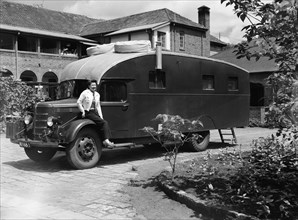 The Price's caravan. A woman sits on the wheel-arch of an old-fashioned caravan in the driveway to