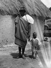 Chief Makimei. Chief Makimei holds the hand of a young boy. He wears a blanket wrapped around one