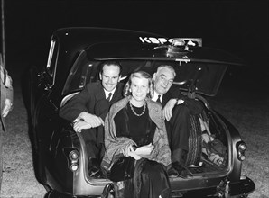 In the boot. Two men wearing suits sit hunched up in the open boot of a car behind a woman wearing
