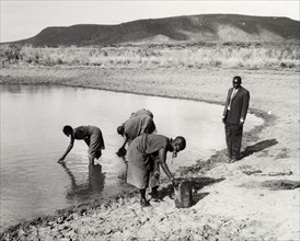 Collecting water in a dry zone. A man in a suit observes as women in traditional dress collect