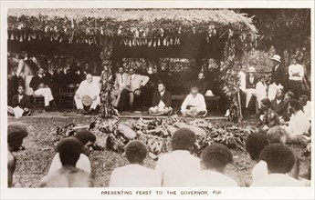 Feast for the Governor, Fiji. An audience of Fijian locals are gathered around a thatched hut,