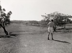 Playing golf, Rangoon. A lone golfer follows his shot, club still swung high in the air, on the