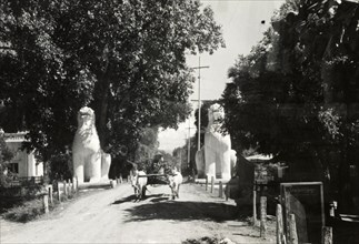 Lion statues guard a Burmese road. A cattle-drawn cart travels along a road guarded by two large,