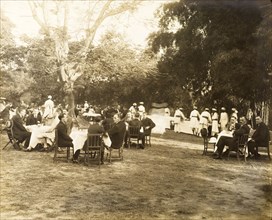Guests at a garden party. Small groups of European men and women sit around dining tables in the