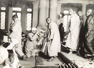Buddhist monks enter a temple. A kneeling Burmese man hands out padded kneelers to a number of