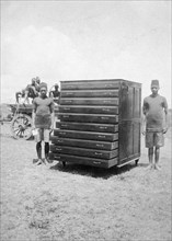 African cabinetmakers. Two young African men stand either side of a large map cabinet they have