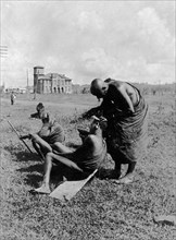 Kikuyu hairdresser. A Kikuyu man dresses the hair of another man seated on a plank on the ground.