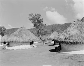 Huts for sugar mill labourers. People seek shade under the thatched roofs of a number of round huts