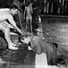 Swimming gala antics. A man in a suit is helped out of a swimming pool by three others wearing
