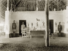 The DC's new office. African colonial officials in uniform stand beside a desk and chairs inside a