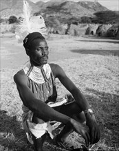 Portrait of a Turkana warrior. A Turkana warrior crouches on the ground. He wears traditional dress