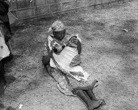 Pepsi cola baby. An African woman sits on the ground, feeding her baby wrapped in a cloth sling