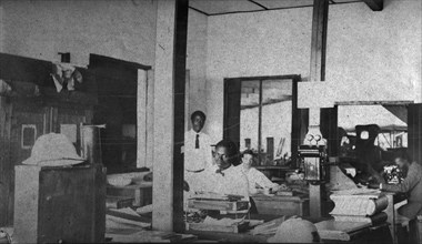 Tamlin in the office. Mr Alfred Tamlin sits in his office accompanied by three African employees.