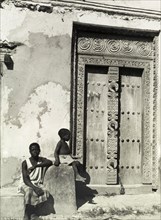 Carved wooden door, Zanzibar. A young man and a child sit in the sunshine beside an ornately carved